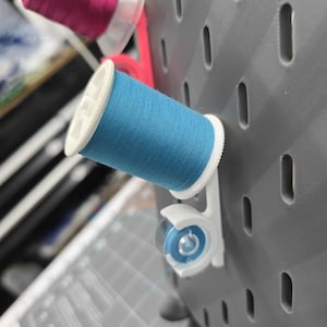 May include: A gray pegboard with several spools of thread in various colors. A blue spool of thread is in the foreground, held by a white plastic holder. Other spools include pink and blue thread. The pegboard has several oval-shaped holes.