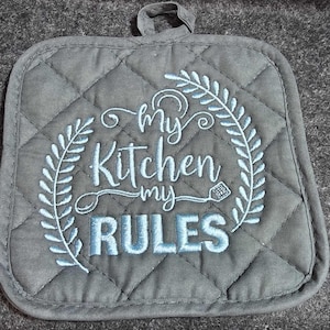 May include: A grey quilted hot pad with a blue embroidered design that says "My Kitchen My Rules" with a wreath of leaves around the text.