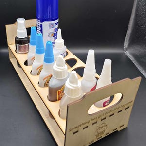 May include: A wooden organizer with eight slots for holding bottles of glue and other adhesives. The organizer has a handle and the text "Something has got to hold it together." printed on the side.