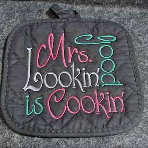 May include: Black quilted pot holder with the embroidered text "Mrs. Lookin' is Cookin'" in pink, grey, and green thread.