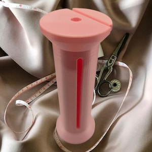 May include: A light pink sewing thread spool holder with a red vertical stripe. A measuring tape, scissors, and a spool of thread are next to the holder. The holder has a circular top with a slot and a hole.