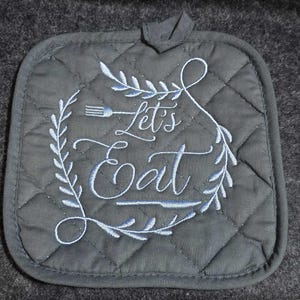May include: A black quilted pot holder with a white embroidered design that says "Let's Eat" with a fork and leaf accents.