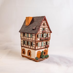 May include: A small, hand-painted ceramic house with a brown roof, white walls, and green shutters. The house has a detailed facade with a wooden frame and flower boxes.
