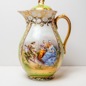 May include: A vintage porcelain creamer with a green and gold design. The creamer features a hand-painted scene of three people in a garden setting.