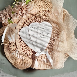 Peut inclure: Un chapeau de paille tressé orné de fleurs et d'éventails d'invitation de mariage. Les éventails sont blancs avec du texte noir, dont "Party" et "Getting Married" avec une date. Les couleurs douces et neutres créent une ambiance romantique.