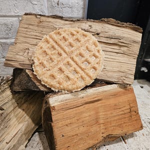 May include: A round, light brown waffle-shaped dog chew sits on top of a stack of wooden logs. The logs are various shades of brown and have a rough, natural texture.