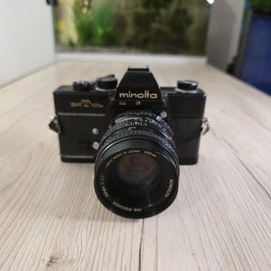 Könnte beinhalten: Eine schwarze Minolta SRT 101b 35mm-Filmkamera mit einem Minolta MD Rokkor 50mm 1:1.7 Objektiv. Das Kameragehäuse hat silberne Akzente, einen Pentaprismensucher und manuelle Einstellräder auf der Oberseite. Der Objektivtubus zeigt den Text Lens Made in Japan und 55mm.