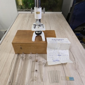 May include: A white and chrome Optico Paris microscope sits atop a wooden storage box. Included are five glass pipettes, several glass slides, and a handwritten document with the header Isly-Optique, detailing the microscope's specifications.