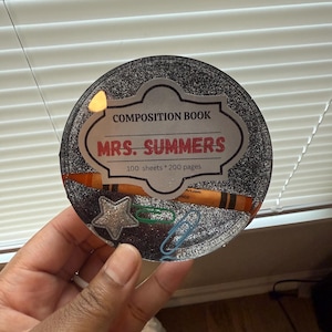 May include: A round, glittery composition book with the text "COMPOSITION BOOK MRS. SUMMERS" and a crayon illustration. Includes a star and paperclip embellishments. The book has 100 sheets and 200 pages.