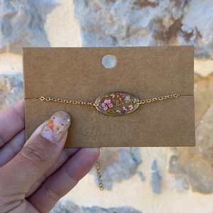 Peut inclure: Bracelet doré avec un pendentif ovale rempli de fleurs séchées roses, violettes et jaunes. Le bracelet est présenté sur une carte marron avec le texte "GACION CREATION". L'arrière-plan est un mur de pierre clair.