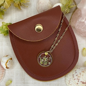 Peut inclure: Une pochette en cuir marron avec une fermeture à bouton-pression dorée contient un collier à chaîne dorée. Le collier présente un pendentif rond transparent avec des fleurs séchées et les lettres "L" et "T". Des coquillages et de la verdure entourent la pochette.