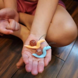 May include: Two handmade clay snails, one light orange and one light blue, each holding a brown, textured sphere. The snails have thin wire antennae. The objects are held in a person's hand.