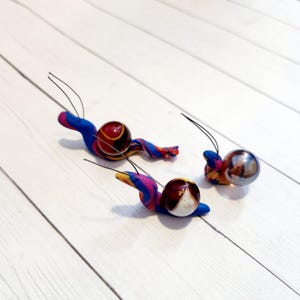 May include: Three handmade snail figurines with colorful, swirled bodies in blue, pink, and yellow. Each snail has a round, decorative shell and thin antennae. The snails are arranged on a white wooden surface.