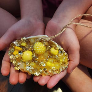 May include: A handcrafted seashell ornament, adorned with yellow beads, sequins, and small yellow decorative elements. The shell is held by hands, and a twine loop is attached for hanging. The ornament has a bright, cheerful appearance.