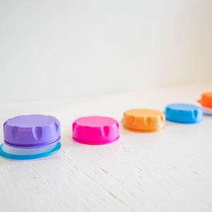 May include: Five colorful plastic lids in a row on a white surface. The lids are purple, pink, orange, blue, and light orange.