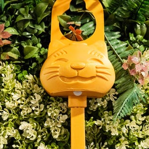 Op de afbeelding: Een feloranje plastic deurhanger met een gestileerd kattengezicht met gesloten ogen en snorharen. Het bovenste gedeelte heeft een ronde uitsparing om op te hangen en een kleine sensor bevindt zich direct onder de kin van de kat.