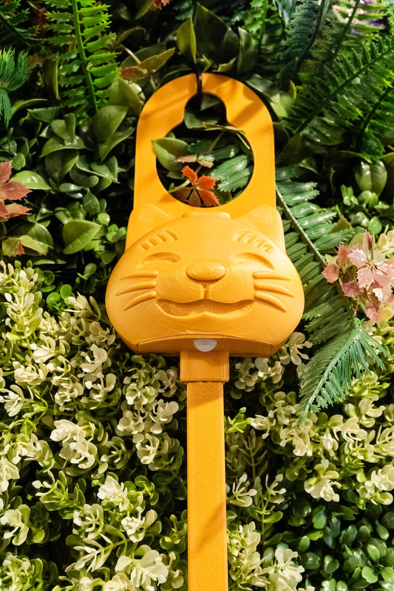 Op de afbeelding: Een feloranje plastic deurhanger met een gestileerd kattengezicht met gesloten ogen en snorharen. Het bovenste gedeelte heeft een ronde uitsparing om op te hangen en een kleine sensor bevindt zich direct onder de kin van de kat.