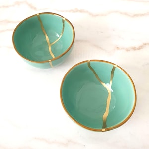 Op de afbeelding: Twee turquoise keramische kommen met goudkleurige kintsugi reparatielijnen. De kommen hebben een afgeronde vorm en een gouden rand. De kintsugi-techniek benadrukt de schoonheid van imperfecties.