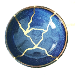 Può includere: Una ciotola in ceramica circolare con un interno smaltato blu intenso e una finitura testurizzata e scintillante. La ciotola è riparata con evidenti linee di lacca dorata che creano un motivo ramificato simile a una ragnatela.