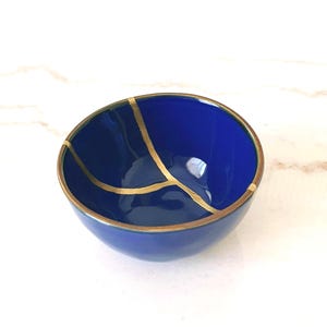 Może przedstawiać: Głęboko niebieska ceramiczna miska ze złotymi liniami i obramowaniem. Miska ma błyszczące wykończenie i unikalny design, inspirowany Kintsugi. Misa stoi na białej powierzchni.
