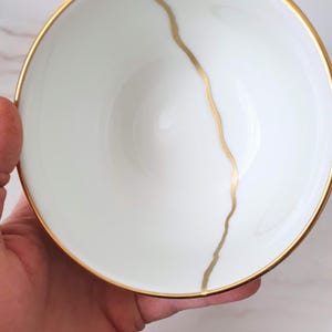 Op de afbeelding: Een witte keramische kom met een gouden rand en een gouden lijn in het midden. De kom wordt in een hand gehouden, wat het delicate ontwerp benadrukt. Perfect voor het serveren van kleine porties of als decoratief item.