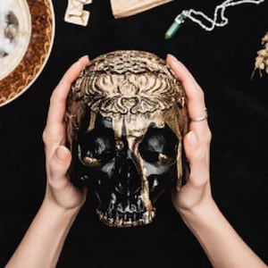 May include: A black and gold decorative skull with ornate detailing on the forehead. The skull is held by two hands against a black background, with other mystical items nearby. The skull is a symbol of the occult.