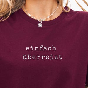 Könnte beinhalten: Ein weinrotes T-Shirt mit Rundhalsausschnitt und dem deutschen Text "einfach überreizt" in Weiß. Das Shirt besteht aus einem weichen Material und wird mit einer cremefarbenen Strickjacke und Jeansshorts getragen. Eine silberne Halskette ist sichtbar.