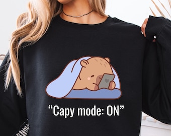 May include: Black long-sleeve sweatshirt featuring a cartoon bear under a blue blanket, holding a phone. The text "Capy mode: ON" is printed below the graphic. The sweatshirt is worn by a person.