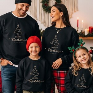 May include: A family wearing matching black sweatshirts with a gold Christmas tree design and the words "MERRY CHRISTMAS AND HAPPY NEW YEAR". The family is standing in front of a Christmas tree and decorations.