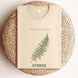 May include: A beige t-shirt with the text "Pflanzen gegen STRESS" and a green fern illustration. The shirt is folded and placed on a woven, circular surface.