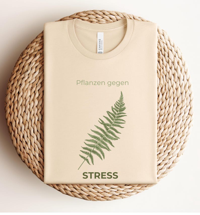 May include: A beige t-shirt with the text "Pflanzen gegen STRESS" and a green fern illustration. The shirt is folded and placed on a woven, circular surface.