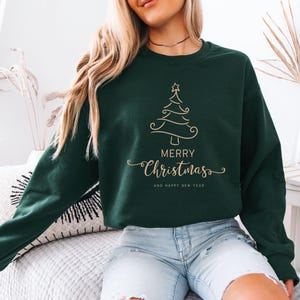 May include: Dark green sweatshirt with a gold Christmas tree design and the words "Merry Christmas" and "And Happy New Year" in a script font. The sweatshirt is a crew neck style.