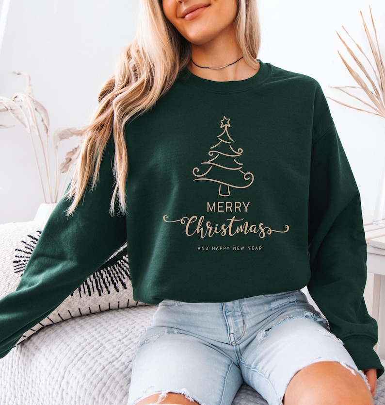 May include: Dark green sweatshirt with a gold Christmas tree design and the words "Merry Christmas" and "And Happy New Year" in a script font. The sweatshirt is a crew neck style.