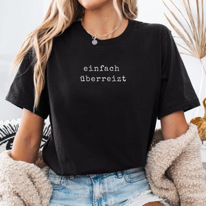 May include: Black t-shirt with the German text "einfach &uuml;berreizt" in white. The shirt is paired with blue denim shorts and a cream-colored jacket. A silver necklace is visible.