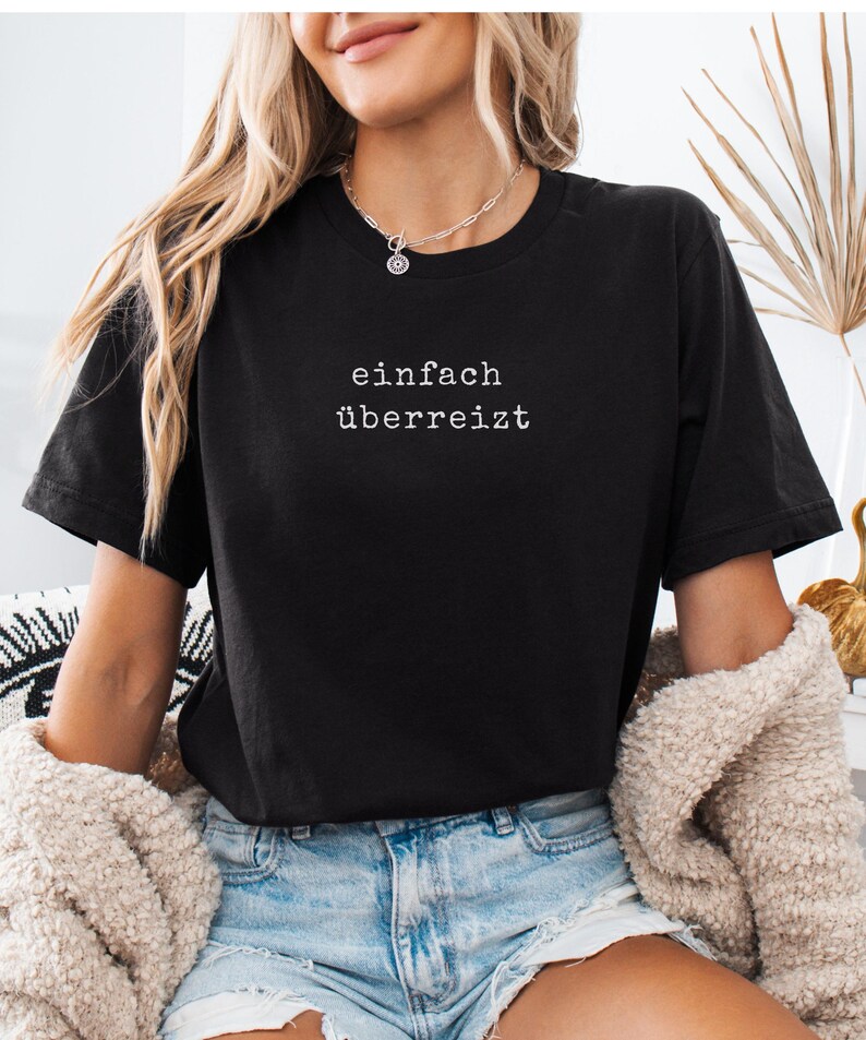 May include: Black t-shirt with the German text "einfach &uuml;berreizt" in white. The shirt is paired with blue denim shorts and a cream-colored jacket. A silver necklace is visible.