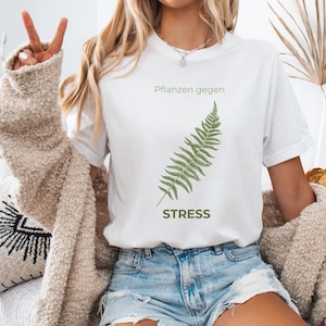 May include: White t-shirt with a green fern illustration and the word "STRESS" in a green font. The text "Pflanzen gegen" is above the fern. The shirt is paired with denim shorts.