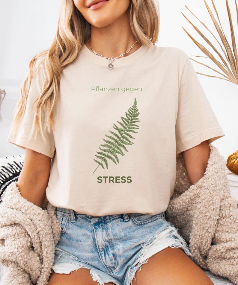 May include: Beige t-shirt with the text "Pflanzen gegen Stress" and a green fern illustration. The shirt is made of a soft material and has short sleeves. The design is centered on the front of the shirt.
