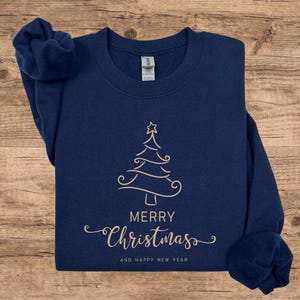 May include: Navy blue sweatshirt with a gold-colored Christmas tree design. The words "MERRY Christmas" and "AND HAPPY NEW YEAR" are printed below the tree. The sweatshirt is folded on a wooden surface.