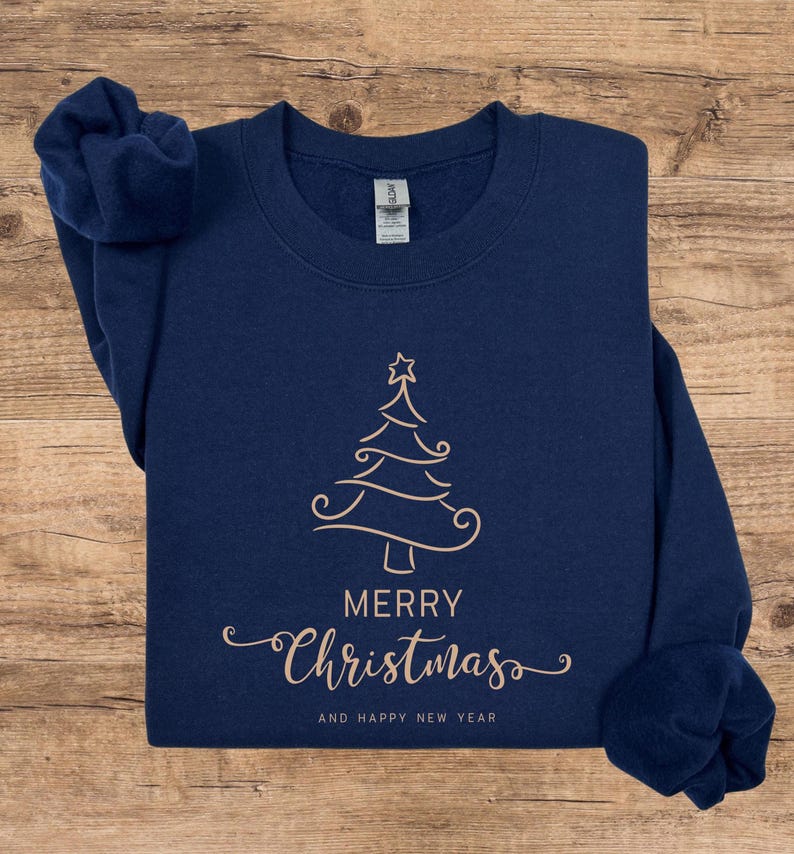May include: Navy blue sweatshirt with a gold-colored Christmas tree design. The words "MERRY Christmas" and "AND HAPPY NEW YEAR" are printed below the tree. The sweatshirt is folded on a wooden surface.