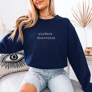 May include: A navy blue sweatshirt with the German text "einfach &uuml;berreizt" in white. The sweatshirt is a long-sleeved crew neck style. The model is wearing light blue jeans.
