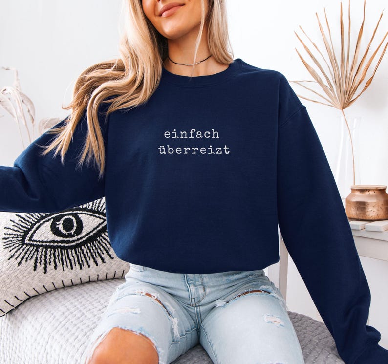 May include: A navy blue sweatshirt with the German text "einfach &uuml;berreizt" in white. The sweatshirt is a long-sleeved crew neck style. The model is wearing light blue jeans.