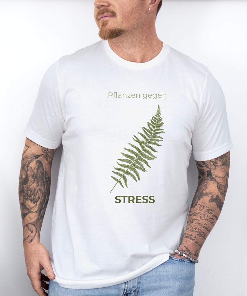 May include: White t-shirt with a green fern illustration and the word "STRESS" below it. The text "Pflanzen gegen" is above the fern. The shirt is made of a soft material.