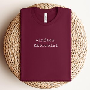 May include: A maroon t-shirt with the words "einfach &uuml;berreizt" printed in white. The shirt is folded and placed on a woven, circular surface. The shirt has a crew neck and a tag.