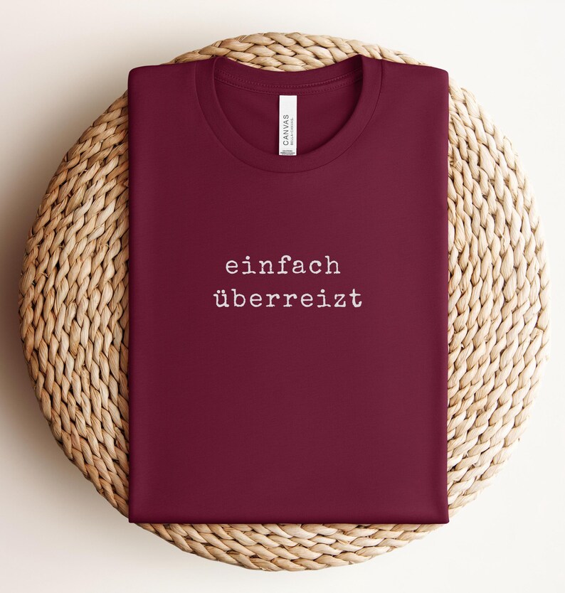 May include: A maroon t-shirt with the words "einfach &uuml;berreizt" printed in white. The shirt is folded and placed on a woven, circular surface. The shirt has a crew neck and a tag.