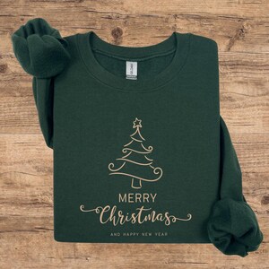 May include: A dark green sweatshirt with a gold-colored Christmas tree design. The words "MERRY Christmas" and "AND HAPPY NEW YEAR" are printed below the tree in a cursive font. The sweatshirt is folded on a wooden surface.