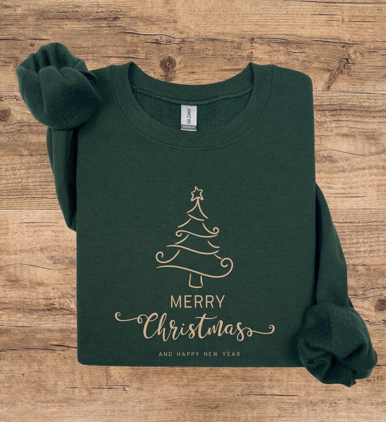 May include: A dark green sweatshirt with a gold-colored Christmas tree design. The words "MERRY Christmas" and "AND HAPPY NEW YEAR" are printed below the tree in a cursive font. The sweatshirt is folded on a wooden surface.