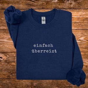 May include: Navy blue sweatshirt with the German text "einfach &uuml;berreizt" in white, centered on the chest. The sweatshirt is made of a soft material and has a crew neck. The sleeves are folded. The background is a wooden surface.
