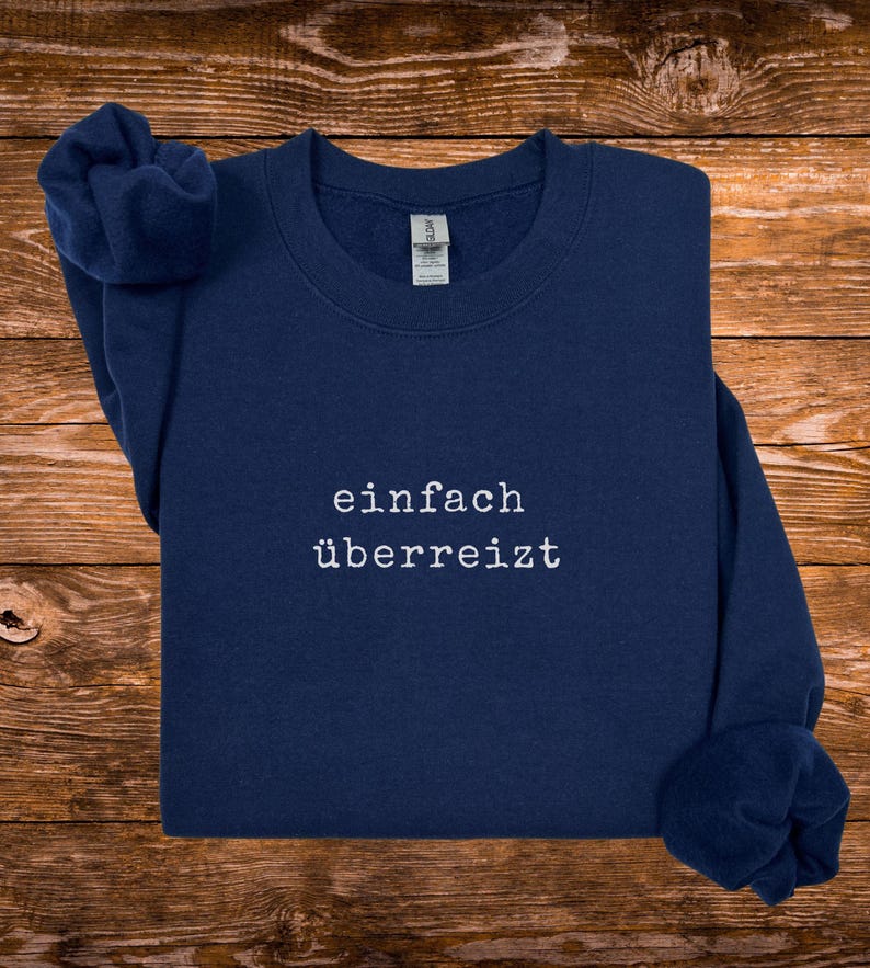 May include: Navy blue sweatshirt with the German text "einfach &uuml;berreizt" in white, centered on the chest. The sweatshirt is made of a soft material and has a crew neck. The sleeves are folded. The background is a wooden surface.