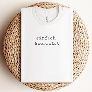 May include: White t-shirt with the German text "einfach &uuml;berreizt" printed in black. The shirt is displayed on a woven, circular surface.