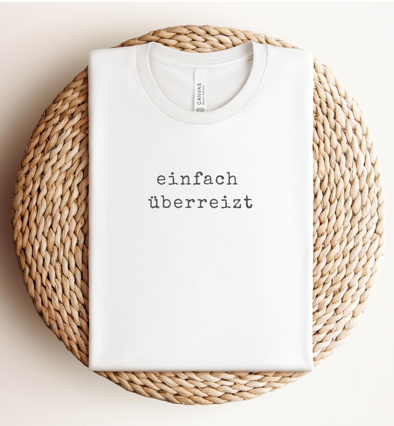 May include: White t-shirt with the German text "einfach &uuml;berreizt" printed in black. The shirt is displayed on a woven, circular surface.